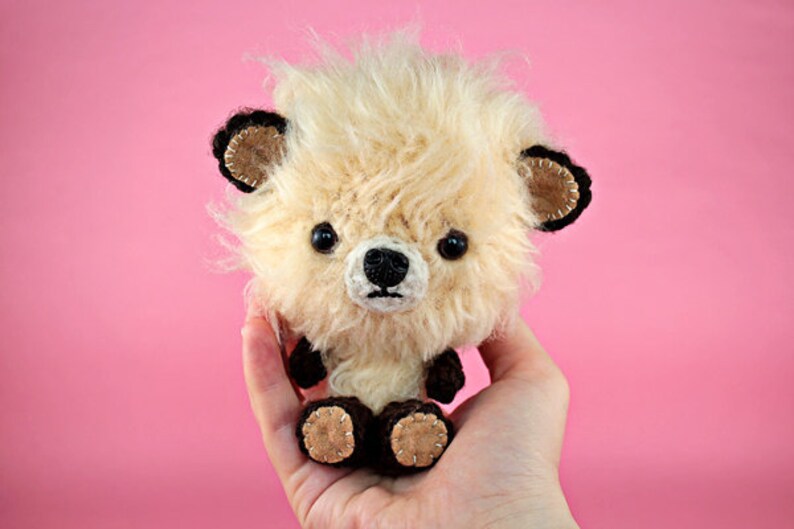 May include: A small, fluffy, cream-colored crocheted stuffed animal with black eyes and a black nose. The animal has brown ears with black trim and brown feet with brown stitching. The animal is being held in a person's hand.