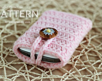May include: A pink crocheted phone case with a wooden button featuring a white daisy. The case is wrapped around a white phone and is lying on a woven surface. The text "Crochet Phone Case PATTERN by zoom yummy.com" is at the top of the image.