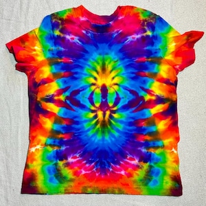 Könnte beinhalten: Ein farbenfrohes Batik-T-Shirt in Rot-, Orange-, Gelb-, Grün-, Blau- und Lilatönen. Das Design weist ein symmetrisches Spiralmuster auf, das von der Mitte ausgeht und einen psychedelischen Effekt erzeugt. Das kurzärmelige T-Shirt ist aus weichem Stoff.