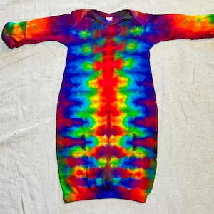 May include: A tie-dye baby gown in a vibrant rainbow of colors, including red, orange, yellow, green, blue, and purple. The gown has long sleeves and a round neckline. The fabric appears to be soft and comfortable.