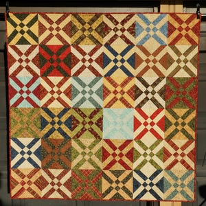 Könnte beinhalten: Ein quadratischer Patchwork-Quilt mit einem sich wiederholenden Blockmuster aus sich kreuzenden diagonalen Linien. Der Quilt besteht aus verschiedenen Stoffquadraten in Rot-, Grün-, Blau-, Gelb- und Cremetönen mit einem dünnen roten Rand.