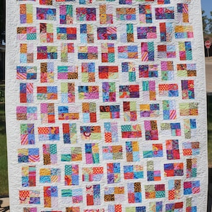 May include: A rectangular quilt featuring a grid of multi-coloured fabric blocks in various patterns, including stripes, dots, and floral prints. The blocks are separated by a white fabric background with a continuous swirling quilted texture.
