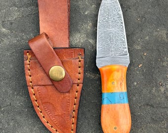 May include: A handmade knife with a patterned blade and an orange handle with a blue stripe. The knife is paired with a brown leather sheath with a belt loop and snap closure. The knife and sheath are displayed on a textured surface.