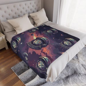 Könnte beinhalten: Eine weiche Decke mit einem sich wiederholenden Muster von Katzen mit Astronautenhelmen vor dem Hintergrund eines lila-orangenen Nebels. Die Decke liegt auf einem Bett mit weißer Bettwäsche und Kissen.