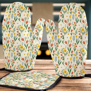 May include: A cream-coloured kitchen set with a floral design. The set includes two oven gloves, a pot holder, and a matching oven glove. The pattern features yellow, orange, and blue flowers on a cream background.