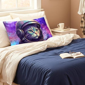 Op de afbeelding: Een rechthoekig kussen met een kat in een astronautenhelm tegen een paarse en blauwe galaxy-achtergrond. De kat heeft groene ogen en is het centrale ontwerpelement. Het kussen ligt op een bed met een blauw dekbed.