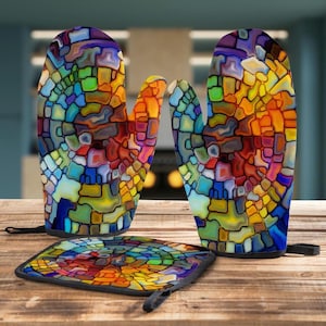 May include: A colorful stained-glass pattern adorns a set of kitchen oven mitts and a matching pot holder. The mitts and pot holder feature a vibrant mosaic of blues, yellows, oranges, and reds, with black trim and hanging loops. The kitchen set is on a wooden surface.