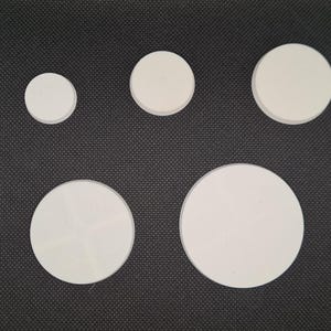 May include: Five white, circular coasters of varying sizes are arranged on a dark gray textured surface. The coasters range from small to large, showcasing a simple, minimalist design. The coasters are made of a light-colored material.