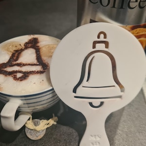 Könnte beinhalten: Ein Cappuccino mit einem Glockenmuster im Schaum, neben einer weißen Glockenschablone. Die Tasse hat blaue Streifen und einen weißen Henkel. Im Hintergrund ist ein Kaffeebehälter zu sehen.