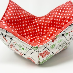 May include: A square fabric bowl with a red polka dot interior and a white exterior patterned with black, red, and green Asian-inspired designs. The bowl is folded to create a unique shape.