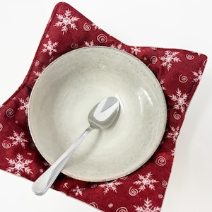 May include: A light grey ceramic bowl with a silver spoon sits on a red, snowflake-patterned bowl cosy. The cosy is square-shaped with white snowflakes and swirls. The spoon is centered in the bowl.