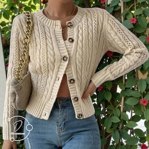 Może przedstawiać: Kremowy kardigan z warkoczowym splotem z brązowymi guzikami. Kardigan ma okrągły dekolt i długie rękawy. Pod spodem widoczny jest pasujący kremowy top. Strój jest połączony z niebieskimi dżinsami i beżową torebką ze złotym łańcuszkiem.