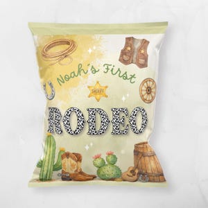 May include: A light green bag with a watercolour design featuring a cowboy theme. The bag has the text "Noah's First Rodeo" with a sheriff's badge, a horseshoe, a cowboy vest, a wagon wheel, a cactus, and a guitar.