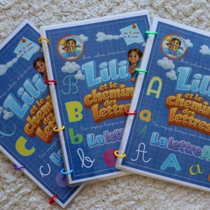 Peut inclure: Trois cahiers d'exercices bleus intitulés Lili et le chemin des lettres, avec des personnages de dessin animé, des lettres de l'alphabet et des quadrillages. Les livrets sont reliés par des anneaux en plastique colorés et un logo rouge.