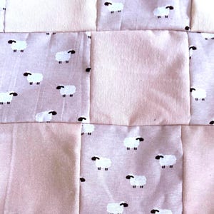 May include: A patchwork quilt featuring alternating square panels of solid light pink fabric and a patterned fabric in a muted mauve tone. The patterned squares are decorated with a repeating print of small white sheep with black heads and legs.