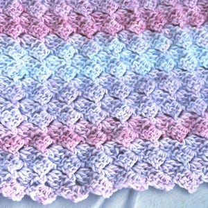 May include: Close-up of a handmade crochet blanket with a textured pattern. The blanket features a gradient of colors, transitioning from pink to light blue and lavender. The edges have a scalloped design.