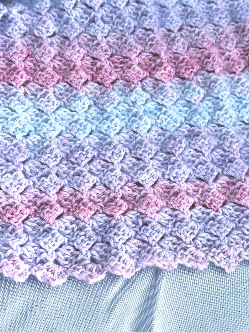 May include: Close-up of a handmade crochet blanket with a textured pattern. The blanket features a gradient of colors, transitioning from pink to light blue and lavender. The edges have a scalloped design.