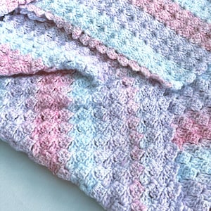 May include: A handmade crochet blanket in soft pastel colors. The blanket features a textured stitch pattern with shades of pink, blue, and lavender. The edge is finished with a scalloped border, adding a delicate touch to the cozy design.