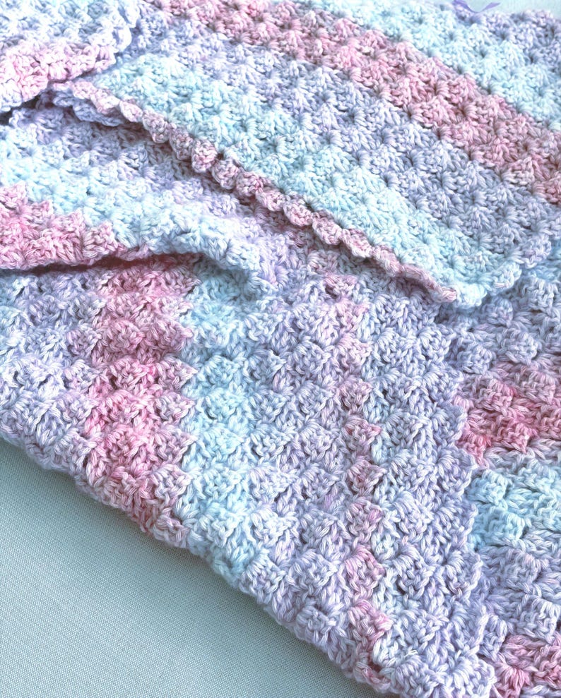 May include: A handmade crochet blanket in soft pastel colors. The blanket features a textured stitch pattern with shades of pink, blue, and lavender. The edge is finished with a scalloped border, adding a delicate touch to the cozy design.