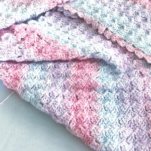 May include: Close-up of a handmade crochet blanket in soft pastel colors. The blanket features a textured stitch pattern with shades of pink, lavender, and light blue. The edges are finished with a scalloped border.