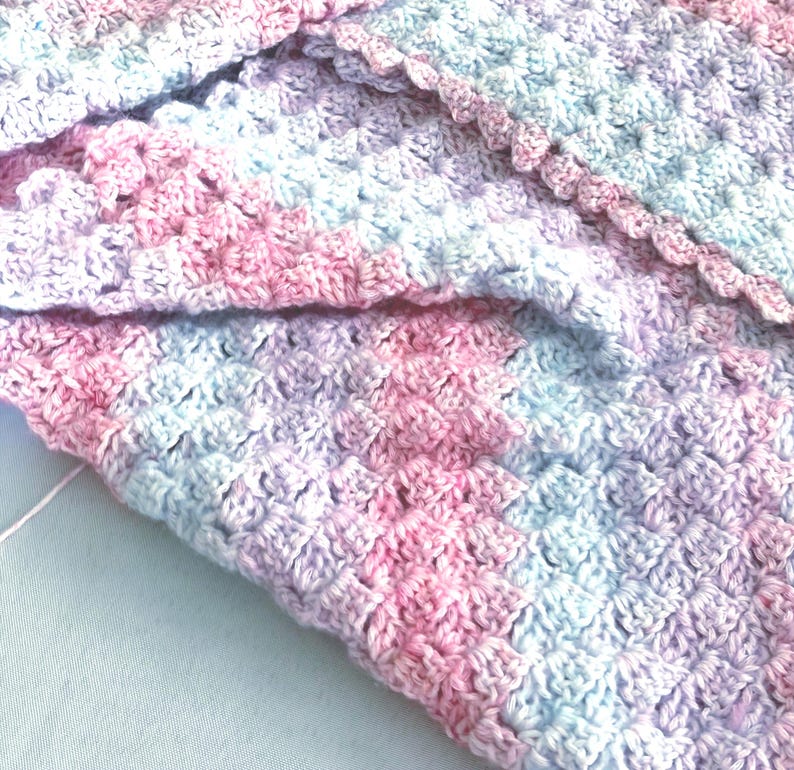 May include: Close-up of a handmade crochet blanket in soft pastel colors. The blanket features a textured stitch pattern with shades of pink, lavender, and light blue. The edges are finished with a scalloped border.