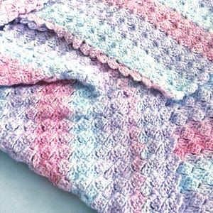 May include: A handmade crochet blanket in soft pastel colors. The blanket features a textured stitch pattern with shades of pink, blue, and lavender. The edges are finished with a scalloped border, adding a delicate touch to the cozy, handcrafted item.