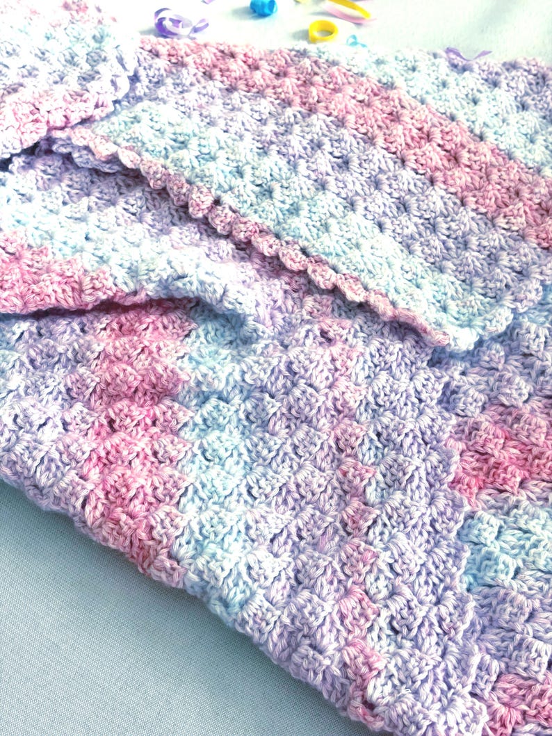 May include: A handmade crochet blanket in soft pastel colors. The blanket features a textured stitch pattern with shades of pink, blue, and lavender. The edges are finished with a scalloped border, adding a delicate touch to the cozy, handcrafted item.