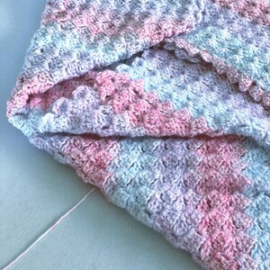 May include: A handmade crochet blanket in soft pastel colors. The blanket features a pattern of pink, blue, and lavender hues. The texture is visible, showcasing the individual stitches. A strand of pink yarn is visible.