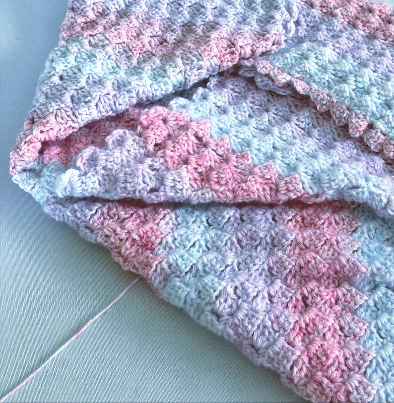 May include: A handmade crochet blanket in soft pastel colors. The blanket features a pattern of pink, blue, and lavender hues. The texture is visible, showcasing the individual stitches. A strand of pink yarn is visible.