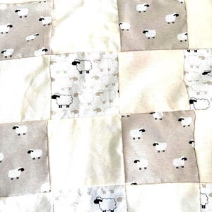 May include: Close-up of a patchwork quilt featuring alternating squares of solid cream fabric and light gray fabric printed with black and white sheep. The quilt has a soft, textured appearance.