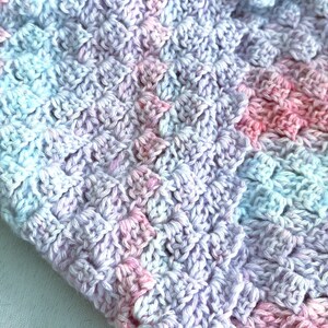 May include: Close-up of a handmade crochet blanket. The blanket features a textured pattern in a blend of pastel colors, including pink, lavender, and light blue. The soft, cozy texture suggests a handmade item, perfect for a baby or as a decorative throw.