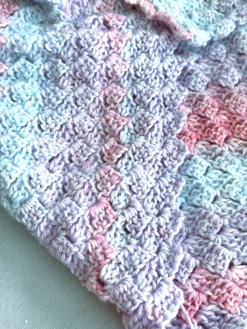 May include: Close-up of a handmade crochet blanket. The blanket features a textured pattern in a blend of pastel colors, including pink, lavender, and light blue. The soft, cozy texture suggests a handmade item, perfect for a baby or as a decorative throw.