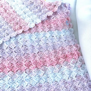 May include: A handmade crochet blanket in soft pastel colors. The blanket features a textured pattern with shades of pink, white, and lavender. The edges are scalloped, adding a delicate touch to the overall design. This cozy blanket is perfect for a baby.