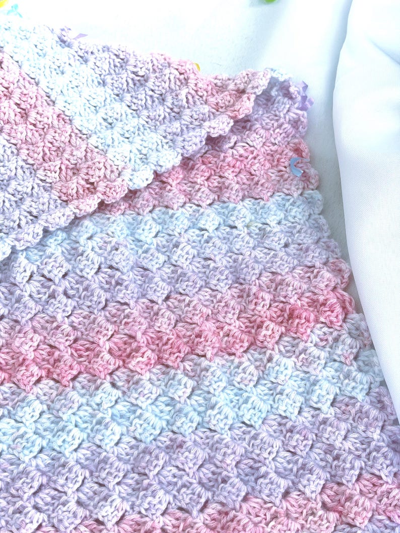May include: A handmade crochet blanket in soft pastel colors. The blanket features a textured pattern with shades of pink, white, and lavender. The edges are scalloped, adding a delicate touch to the overall design. This cozy blanket is perfect for a baby.