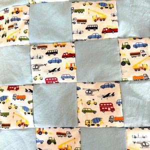 May include: A patchwork quilt featuring an alternating grid pattern of solid light blue squares and white squares printed with a colorful variety of vehicles including red buses, yellow construction trucks, green tractors, and airplanes.