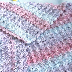 May include: A handmade crochet blanket in soft pastel colors. The blanket features a textured stitch pattern with shades of pink, lavender, and light blue. The edges are finished with a scalloped border, adding a delicate touch to the cozy, handmade item.