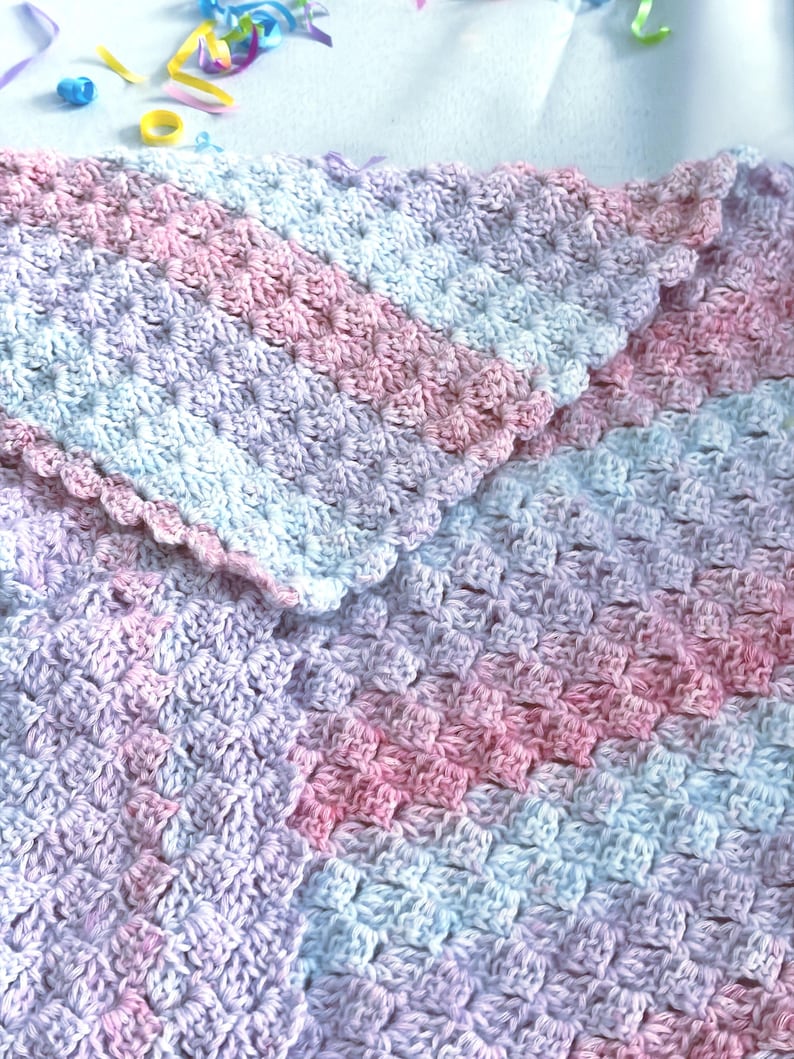 May include: A handmade crochet blanket in soft pastel colors. The blanket features a textured stitch pattern with shades of pink, lavender, and light blue. The edges are finished with a scalloped border, adding a delicate touch to the cozy, handmade item.