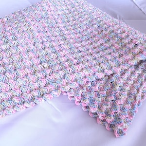 May include: A handmade crochet blanket in pastel colors. The blanket features a textured pattern with pink, blue, and white yarn. The blanket is folded, showcasing the detailed stitch work and soft color palette.