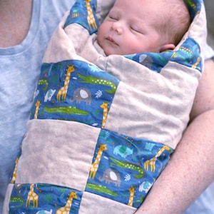 May include: A sleeping baby wrapped in a patchwork baby blanket. The blanket features a blue fabric with a pattern of giraffes, elephants, and crocodiles. The blanket is beige and blue.