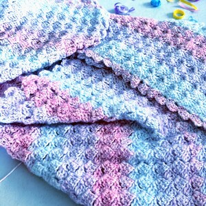 May include: A handmade crochet blanket in shades of blue, pink, and lavender. The blanket features a textured stitch pattern and a scalloped edge. The soft color palette and cozy texture suggest a baby blanket or lap throw.