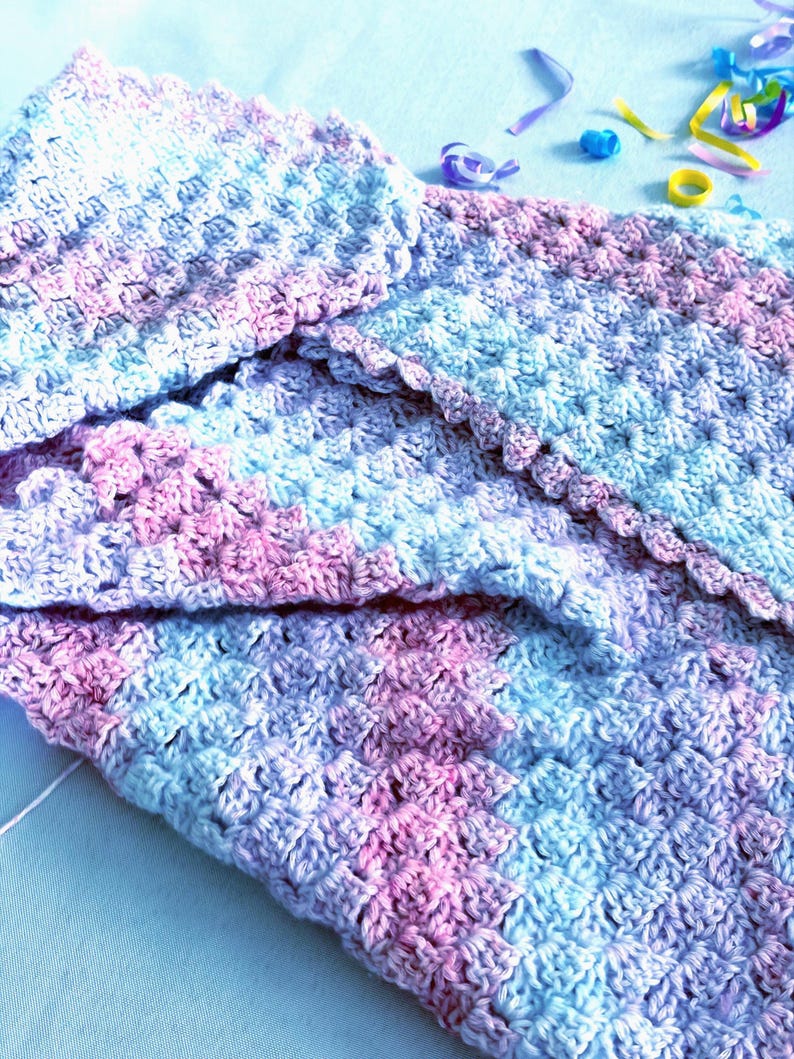 May include: A handmade crochet blanket in shades of blue, pink, and lavender. The blanket features a textured stitch pattern and a scalloped edge. The soft color palette and cozy texture suggest a baby blanket or lap throw.