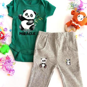 May include: A green baby bodysuit with a panda bear graphic and the text "tiny miracle". Gray baby pants with two panda bear graphics. An orange crochet rattle with a wooden ring is also present.