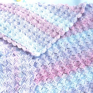 May include: A handmade crochet blanket in shades of pink, white, and lavender. The blanket features a textured stitch pattern and a scalloped edge. The soft color palette and cozy design suggest a baby blanket or lap throw.