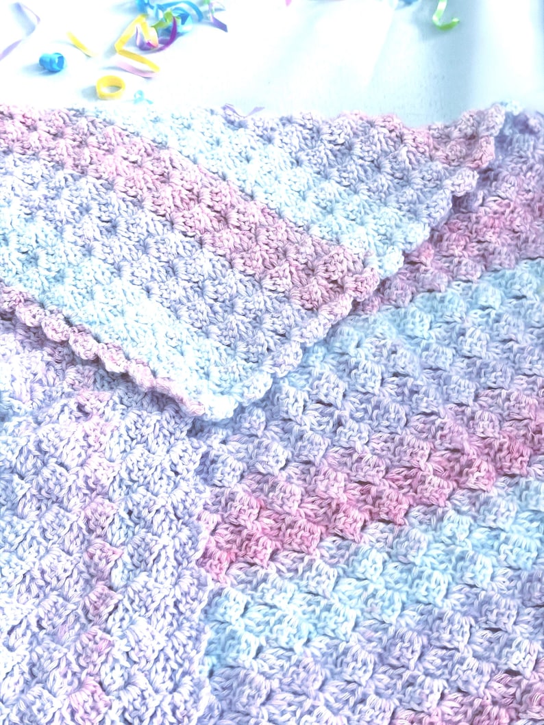 May include: A handmade crochet blanket in shades of pink, white, and lavender. The blanket features a textured stitch pattern and a scalloped edge. The soft color palette and cozy design suggest a baby blanket or lap throw.