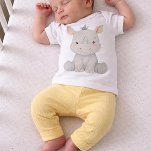 May include: A sleeping baby wearing a white onesie with a rhino graphic and yellow pants. The baby is lying on a white crib sheet with a dotted pattern. The onesie features a smiling rhino.