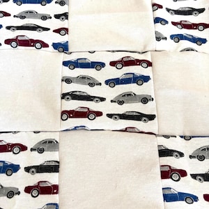 May include: A patchwork quilt featuring alternating squares of solid off-white fabric and a patterned fabric printed with rows of vintage sports cars in red, blue, black, and gray. The design consists of a repeating grid of these fabric blocks.