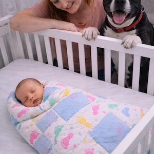 May include: A baby swaddled in a blue, white, and pastel-colored foot-print patterned quilt, resting in a white crib. A woman and a black and white dog are looking over the crib.