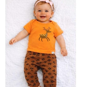 May include: A baby wearing an orange t-shirt with a deer design and brown trousers with a paw print pattern. The baby has a pink bow headband.