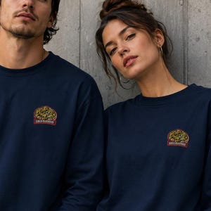 Könnte beinhalten: Ein dunkelblaues Langarmshirt mit einer kleinen, kreisförmigen Grafik auf der linken Brust. Die Grafik zeigt ein grünes Reptil mit dem darunter in Rot gedruckten Text COLD BLOODED. Das Shirt hat einen Rundhalsausschnitt und ein schlichtes Design.