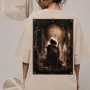 Könnte beinhalten: Beiges T-Shirt mit großem Aufdruck auf dem Rücken, der eine Figur in einer dunklen Kathedrale zeigt, mit dem Text "SHE IS THE SHIELD" und "VOID CULT". Die Vorderseite des Shirts hat ein kleineres Logo mit einem Kreuz.